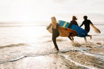 Вид сбоку на группу друзей-серферов, одетых в гидрокостюмы, бегущих с досками для серфинга к воде, чтобы поймать волну на пляже во время восхода солнца — стоковое фото