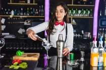 Female barkeeper in stylish outfit adding ice cubes into a glass while preparing cocktail standing at counter in modern bar — Stock Photo