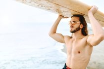 Homem surfista jovem com cabelo comprido vestido de fato de mergulho e óculos de sol elegantes de pé carregando a prancha de surf na cabeça olhando para longe na praia — Fotografia de Stock