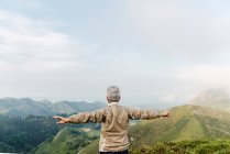 Обратный вид на неузнаваемую старшую женщину-путешественницу, поднимающую и вытягивающую руки утром на холмистой местности — стоковое фото