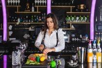 Young female barkeeper in stylish outfit preparing mojito cocktail with lemon slices while standing at counter in modern bar — Stock Photo