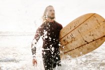 Hombre surfista vestido con traje de neopreno corriendo con tabla de surf en la playa durante el amanecer - foto de stock