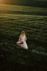 Молода жінка в вінтажній сукні з плетеним кошиком під час прогулянки в зеленому трав'янистому полі під час заходу сонця в літній сільській місцевості — стокове фото