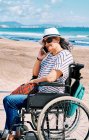 Vista lateral do viajante feminino deficiente positivo sentado em cadeira de rodas e falando no smartphone enquanto passa férias de verão na praia perto do oceano ondulante — Fotografia de Stock