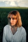 Мирная молодая женщина в белой блузке в стиле ретро сидит посреди высокой зеленой травы и смотрит в камеру во время летнего отдыха в сельской местности — стоковое фото