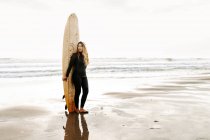 Surferin im Neoprenanzug steht mit dem Surfbrett am Strand bei Sonnenaufgang im Hintergrund und schaut weg — Stockfoto