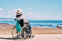 Вид ззаду на невпізнавану жінку-мандрівника в інвалідному візку з рюкзаком, насолоджуючись літньою подорожжю на пляжі біля блакитного моря — стокове фото
