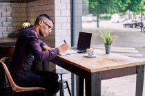 Серьезный латиноамериканец-менеджер в фиолетовой рубашке и очках, печатающий на нетбуке, сидя за столом за окном и работая над проектом в кафе — стоковое фото