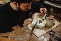 Бородатий чоловічий золотошукач використовує щипці, роблячи крихітні металеві деталі на верстаті в майстерні — стокове фото