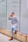 Homem hispânico de corpo inteiro em roupa elegante olhando para longe e falando no celular enquanto se inclina na parede na rua da cidade — Fotografia de Stock