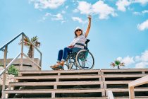 Low angle full body of positive handicapped female sitting in wheelchair near stairway and waving hand while looking away against blue sky in tropical city — Stock Photo
