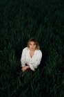 Спокійна молода жінка в ретро-стилі біла блузка сидить серед високої зеленої трави і дивиться на камеру, відпочиваючи в літній вечір у сільській місцевості — стокове фото