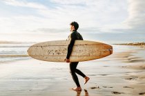 Vista laterale dell'uomo surfista vestito di muta che cammina con la tavola da surf verso l'acqua per catturare un'onda sulla spiaggia durante l'alba — Foto stock