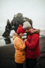 Side view of young man and woman in winter wear standing on coast near big stone and water — Stock Photo