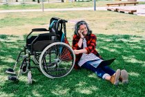 Повне тіло позитивної сірої волохатої жінки з ноутбуком на колінах, що відповідає на телефонний дзвінок, сидячи на зеленій траві біля інвалідного візка в парку — стокове фото