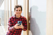 Молодой мужчина в повседневной одежде с помощью смартфона, стоя перед камерой в современном городском здании — стоковое фото