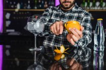 Cropped unrecognizable male barkeeper adding orange peel into a glass while preparing cocktail standing at counter in modern bar — Stock Photo