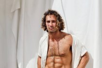 Serious fit shirtless macho com cabelo encaracolado olhando para a câmera no fundo branco no dia ensolarado — Fotografia de Stock