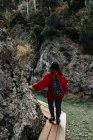 Back view of young tourist with backpack walking on walkway above water near stone wall between forest — Stock Photo