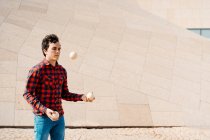 Habilidade jovem macho em camisa quadriculada realizando truque com bolas de malabarismo, enquanto em pé contra a estrutura de concreto contemporâneo na rua urbana — Fotografia de Stock
