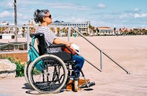Vista laterale viaggiatore femminile in sedia a rotelle con zaino godendo il viaggio estivo sulla spiaggia vicino al mare blu — Foto stock