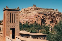 Ancient buildings of brown stone among green tropical plants and desert with clear blue sky on background in Morocco — Stock Photo