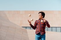 Qualifizierter junger Mann im karierten Hemd, der einen Trick mit Jonglierbällen vollführt, während er auf der städtischen Straße gegen eine moderne Betonkonstruktion steht — Stockfoto