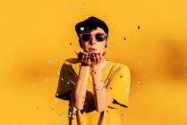 Femme moderne en casquette hip hop et lunettes de soleil soufflant des confettis colorés à la caméra tout en s'amusant sur fond jaune — Photo de stock