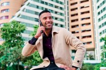Niedriger Winkel positiver gut gekleideter junger hispanischer Geschäftsmann, der auf dem Smartphone spricht und Nachrichten über urbane Straßen diskutiert, mit zeitgenössischen Gebäuden im Hintergrund — Stockfoto