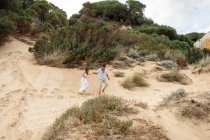 Deleitado puente multirracial y novio tomados de la mano y corriendo a lo largo de la colina de arena en el día de la boda en la naturaleza - foto de stock