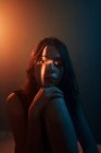 Giovane modello femminile senza emozioni con proiezione di luce a forma di croce sul viso seduto in studio buio e guardando la fotocamera — Foto stock