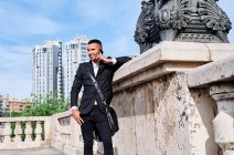 Ganzkörperhispanischer Mann in stylischem Outfit, der wegschaut und mit dem Handy telefoniert, während er sich auf der Stadtstraße an die Wand lehnt — Stockfoto