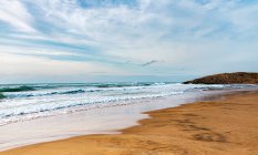 Захватывающий дух вид пенного волнистого океана возле песчаного побережья, расположенного под облачным ярким небом при дневном свете — стоковое фото