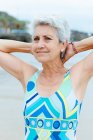 Glücklich gealterte grauhaarige Frau in stylischer heller Badebekleidung hält die Hände hinter dem Kopf, während sie am Strand am Meer Übungen macht — Stockfoto