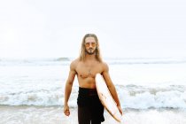 Homem surfista jovem com cabelo comprido vestido de fato de mergulho e óculos de sol elegantes olhando para a câmera com prancha de surf em direção à água para pegar uma onda na praia — Fotografia de Stock
