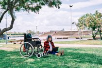Ganzkörper positive grauhaarige Frau mit Laptop auf Knien sitzt auf grünem Gras in der Nähe von Rollstuhl im Park — Stockfoto