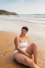 Sorrindo jovem plus size fêmea em maiô sentado na praia de areia olhando para longe perto do oceano espumoso sob céu azul nublado à luz do dia — Fotografia de Stock