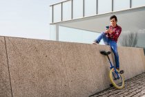 Полное тело молодого парня в клетчатой рубашке и джинсах, сидящего на каменном заборе возле одноколесного велосипеда и просматривающего мобильный телефон в свободное время в городе — стоковое фото