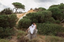 Multiracial couple of newlyweds sitting on stump of tree in woods and hugging on wedding day — Stock Photo