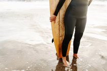 Gestutzte, bis zur Unkenntlichkeit verkleidete Surferin im Neoprenanzug, die mit dem Surfbrett am Strand beim Sonnenaufgang im Hintergrund wegschaut — Stockfoto