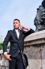 Confident young man in elegant formal suit talking on mobile phone and smiling while standing near sculpture on urban square — Stock Photo