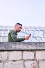 Niedriger Winkel eines erwachsenen hispanischen Mannes im Hemd, der sich an eine Steinmauer lehnt und außerhalb des modernen Glasgebäudes mit dem Handy surft — Stockfoto