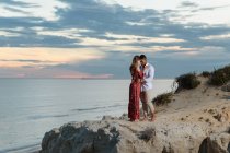 Amando casal multirracial em roupas elegantes abraçando na colina no fundo do céu por do sol sobre o mar no verão — Fotografia de Stock