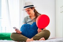 Young female in casual clothes and hat holding red balloon and heart shaped cushion browsing on smartphone while celebrating birthday alone on bed with food and juice — Stock Photo