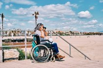 Вид сбоку неузнаваемой волосатой женщины-инвалида в инвалидной коляске, снимающей на фотокамеру, проводя летний день на пляже — стоковое фото