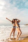 Повне тіло веселих подружок у смугастому купальнику, що стоїть на піщаному пляжі, омитому хвилею моря — стокове фото