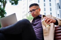 Full length of confident young Hispanic man in stylish casual outfit and glasses answering phone call while sitting on bench and working with laptop on city street — Stock Photo
