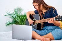 Акцент на просмотре видео-урока для студенток на ноутбуке во время обучения игре на акустической гитаре в свободное время дома — стоковое фото