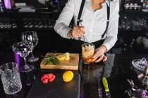 Cropped unrecognizable female barkeeper in stylish outfit adding ice cubes into glass and stirring with long spoon while preparing cocktail standing at counter in modern bar — Stock Photo