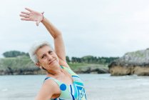 Positive Seniorin im stylischen Badeanzug macht Side-Bend-Übungen, während sie einen gesunden Lebensstil praktiziert und am Strand gegen das wogende Meer trainiert — Stockfoto